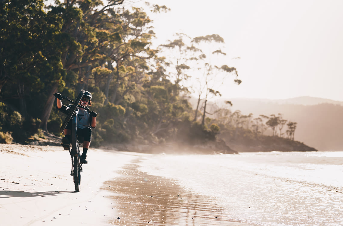 Sascha Kim wheelies a mountain bike along the beach.