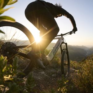 A mountain biker rides into a descent on a Giant Stance E+ with the sun setting in the background.