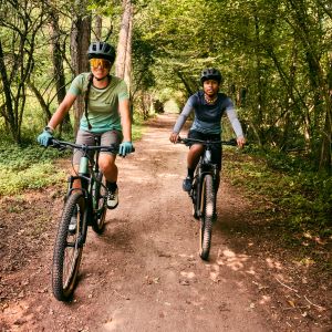 Two women riding Liv Tempt E+ hardtail mountain bikes through a wooded trail.