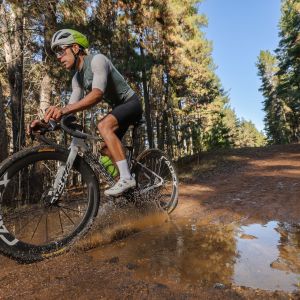 A gravel cyclist on a Giant Revolt Advanced SL charges through a puddle on a dirt road wearing Giant Charge Pro gravel/XC shoes.