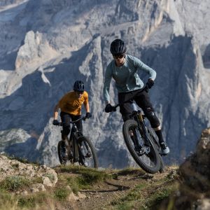 Two mountain bikers on Giant Trance Advanced E+ trail bikes with Giant TRX wheels ride over the crest of a rocky mountain trail.