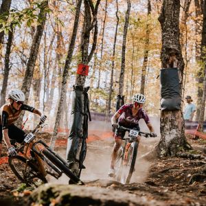 A CX mountain biker races through a technical section on her Liv Pique Advanced SL past another rider.