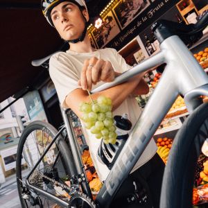 A cyclist carries their Giant Contend SL aluminum road bike through a small market with a handful of grapes.