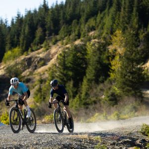 Two cyclists riding Giant Revolt Advanced SL gravel bikes navigate a dirt mountain road around a lake at speed.