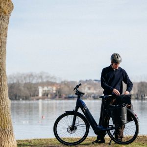 A cyclist stands next to their Giant NewTour E+ city bike on the edge of a body of water while going through their rear pack.
