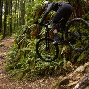 A mountain biker hits a medium sized drop on a forest trail riding a Giant Stance.