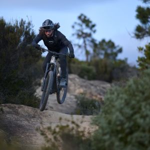 A mountain biker lands a small jump on a down-hill trail on a Giant Stance E+ trail bike.