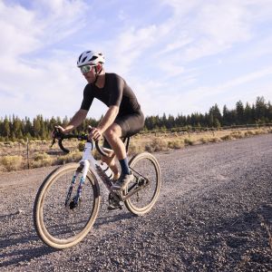 A gravel cyclist rides a dirt road with pine trees in the background on their Giant Revolt Advanced SE.