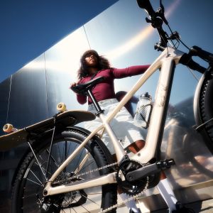 A female cyclist leans against a building next to her bike with her skateboard strapped to the rack.