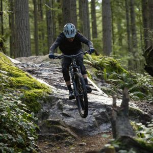 A mountain biker riding a Giant Stance trail bike hits a technical rocky drop on a forest trail.