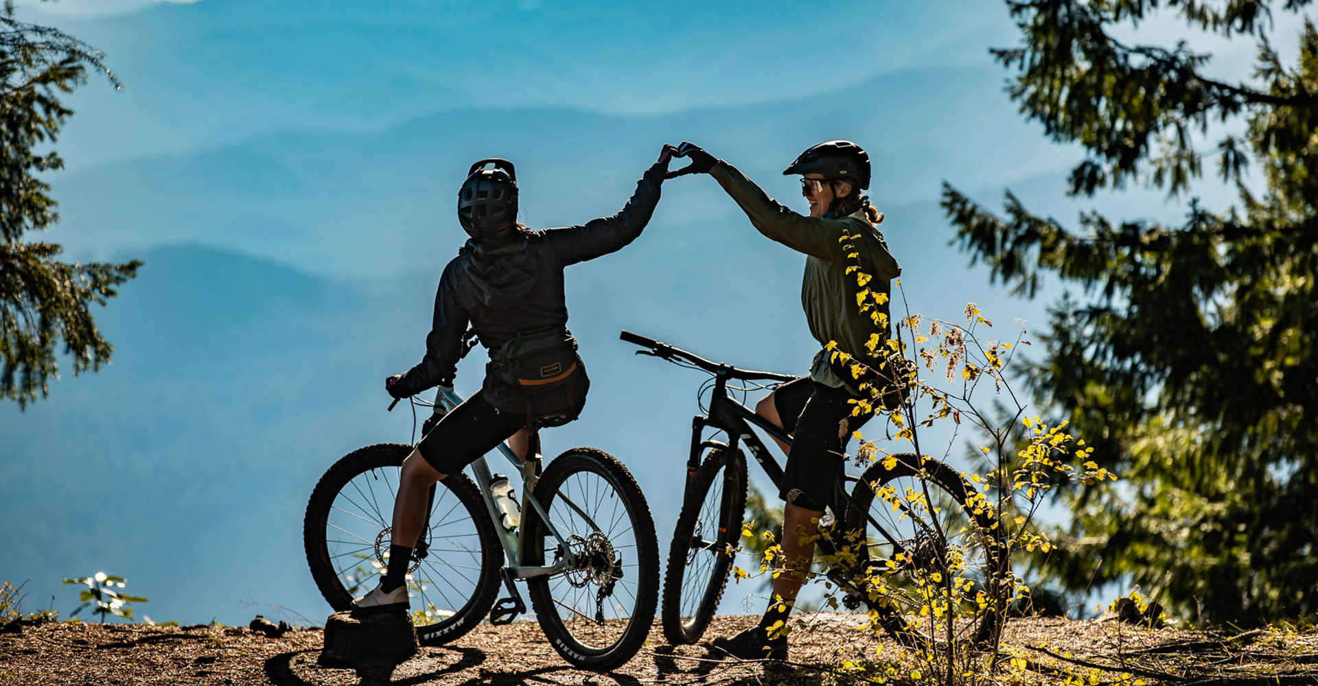 Two women mountain bikers stopped at a viewpoint, using their hands together to make a heart